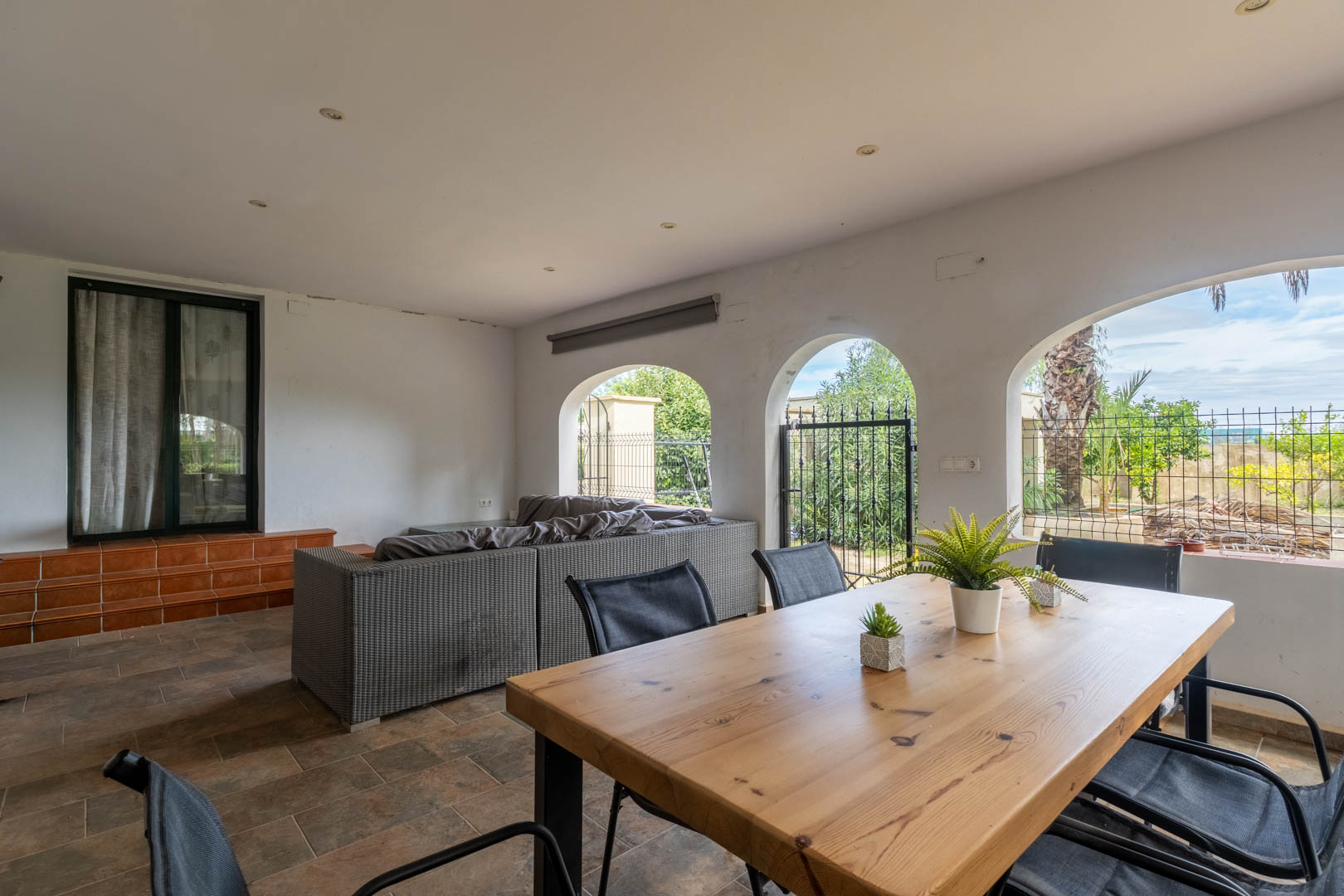 Covered terrace featuring Mediterranean arches, a solid wood dining table, rattan sofas, and garden views. Rustic tiled flooring and recessed ceiling lighting.