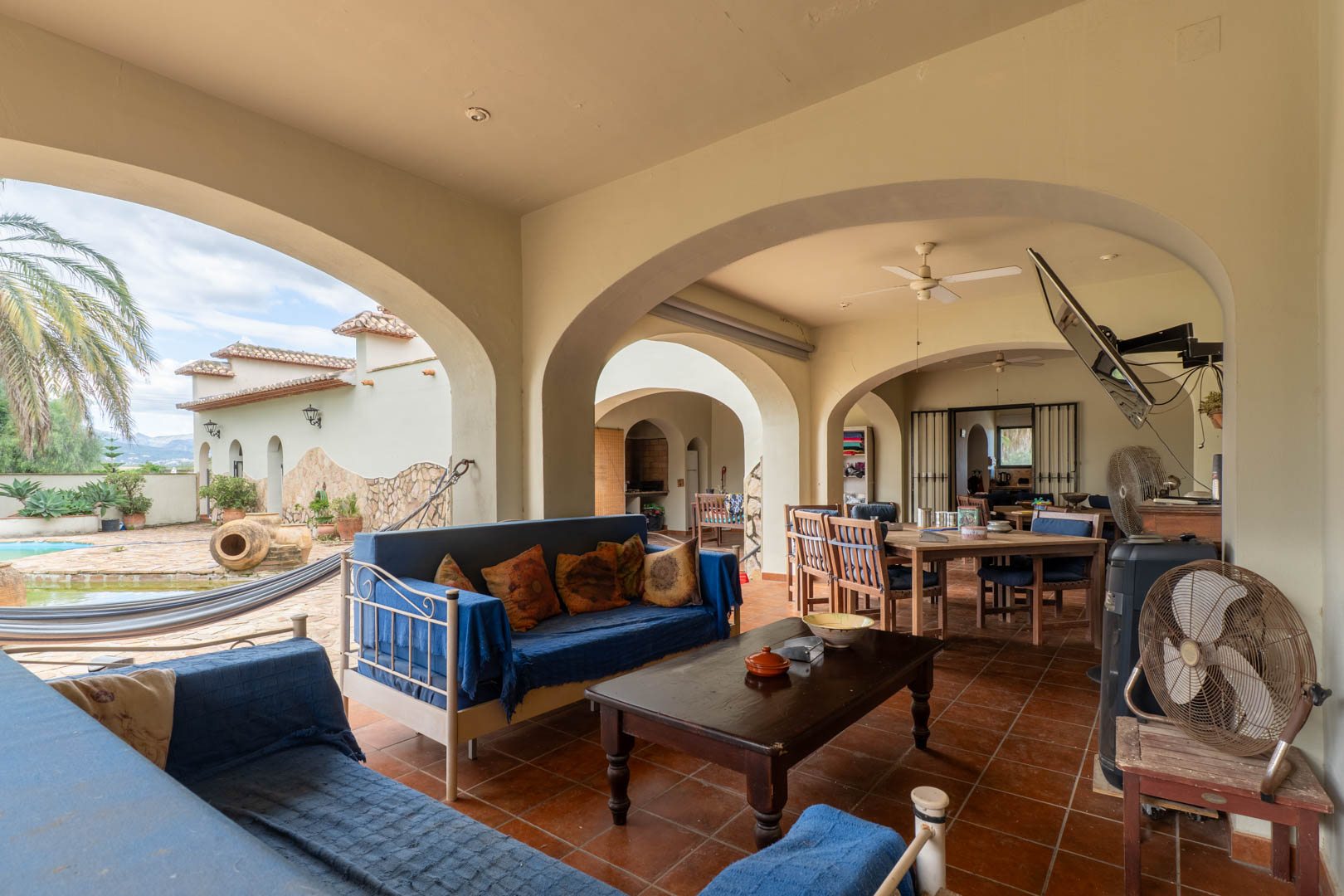Covered terrace featuring Mediterranean arches, terracotta flooring, and a lounge area. It overlooks the swimming pool, hammock, and gardens with mature palm trees.