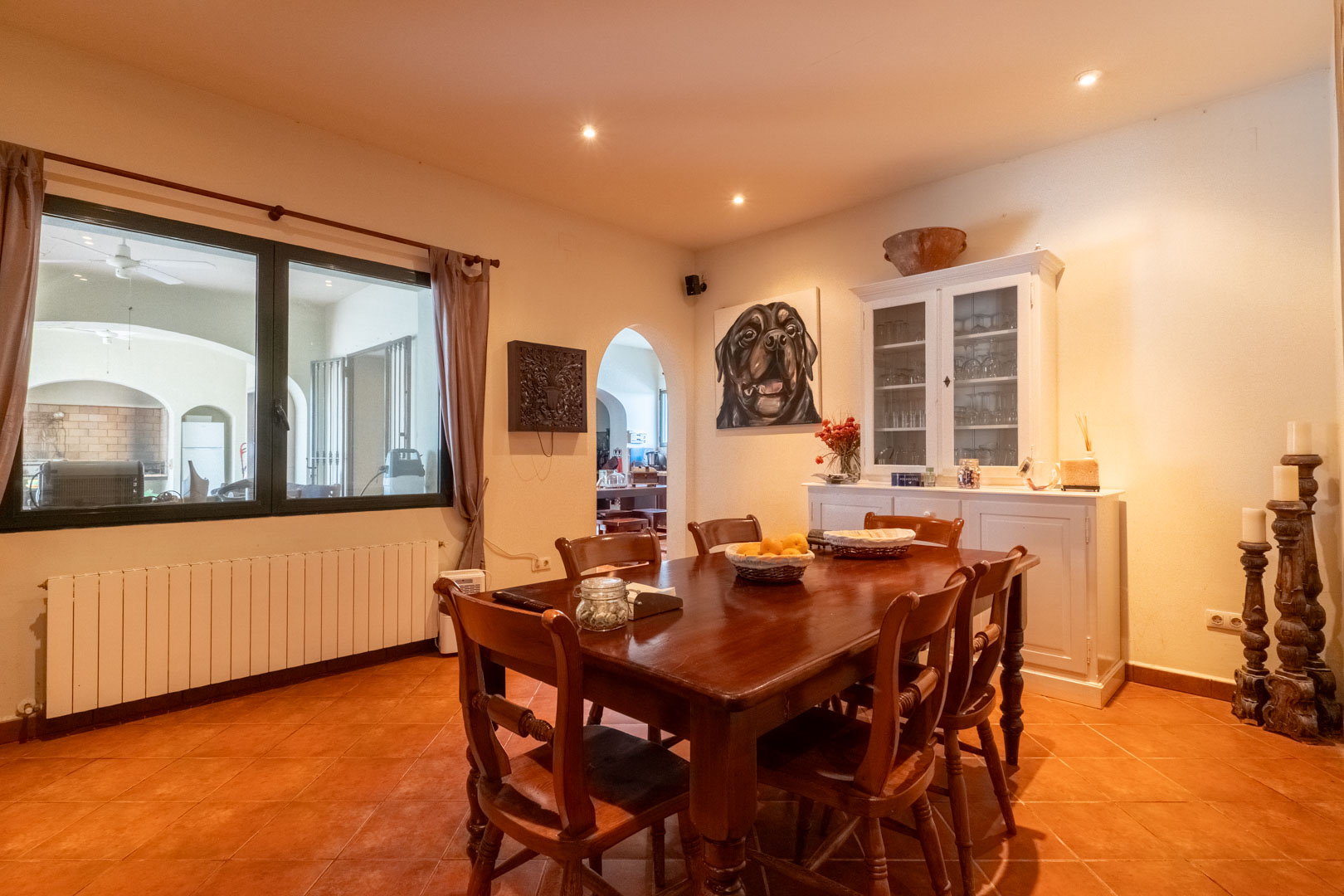 Dining area featuring terracotta flooring, a solid wood table set, and a white sideboard. A large window overlooks the porch, complemented by recessed lighting.
