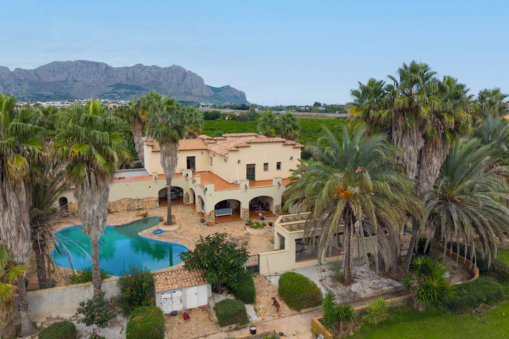 Elevated view of the Mediterranean estate showing the freeform pool, arched terraces, and extensive palm groves with panoramic mountain views.