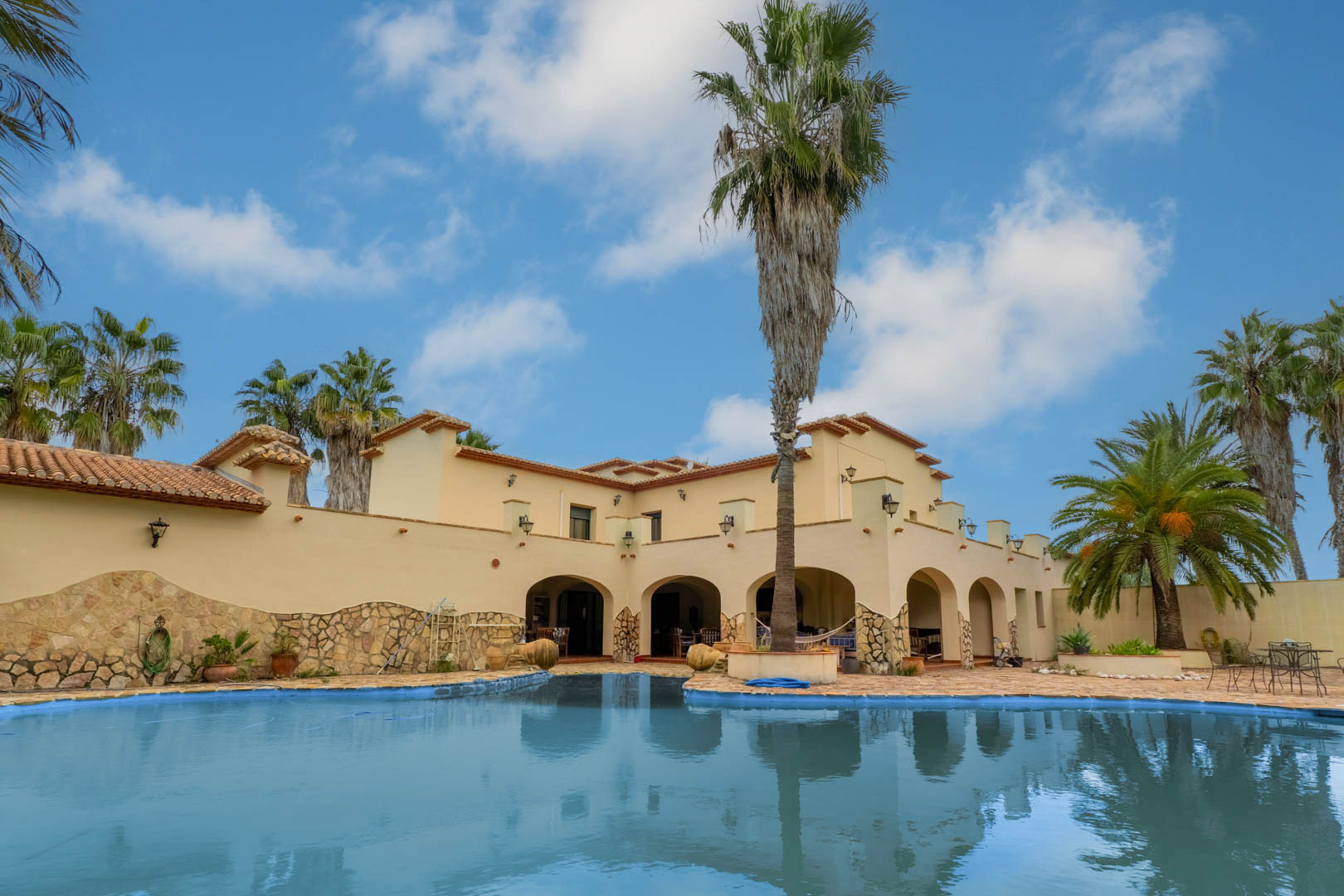 Exterior view of the irregular-shaped swimming pool and stone terrace, featuring the villa's Mediterranean facade with arched porches and tall palm trees.