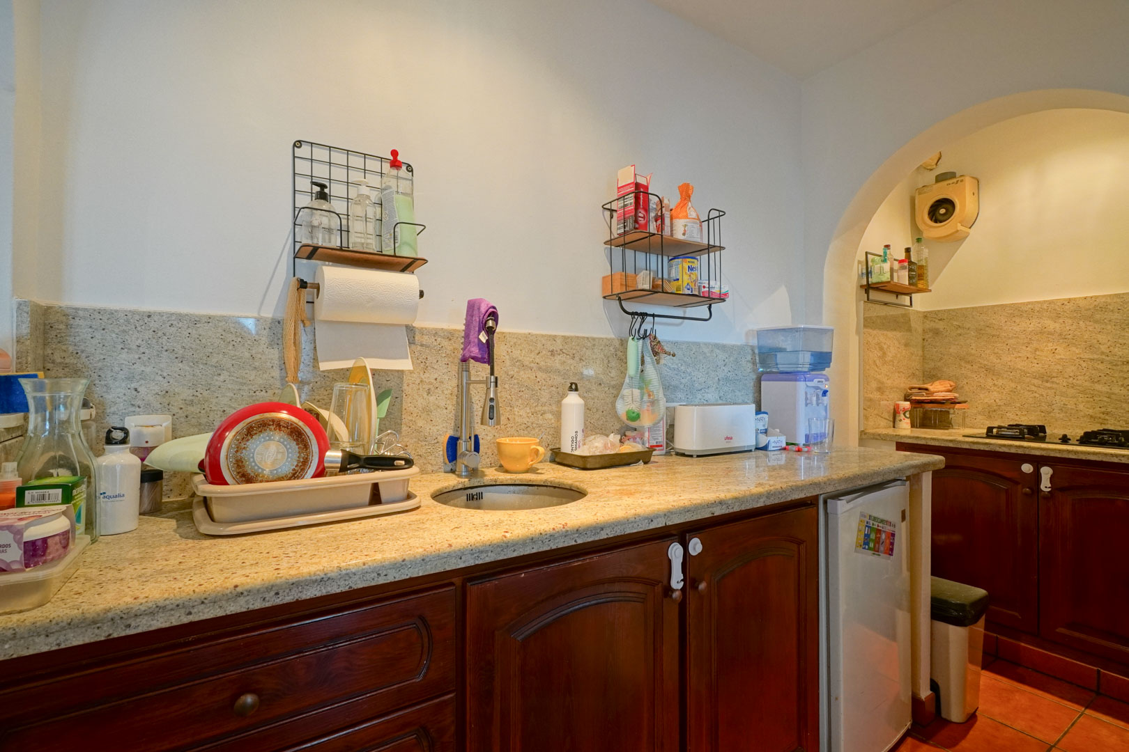 Functional kitchen featuring granite countertops, dark wood cabinetry, and a circular sink, with an arched opening to the cooking area.