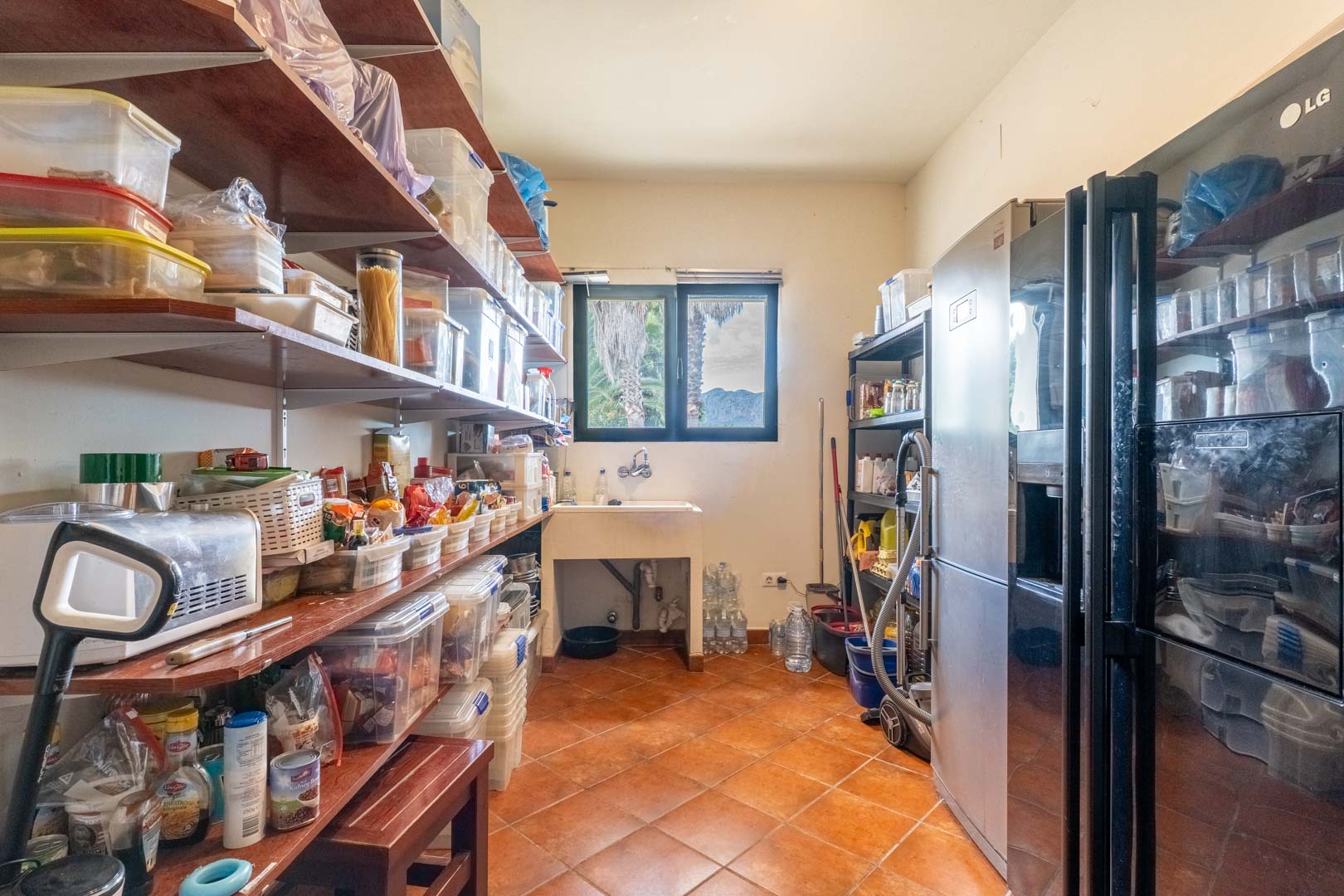 Functional pantry featuring extensive wooden shelving, a utility sink under a window with mountain views, and large refrigeration units on tiled flooring.
