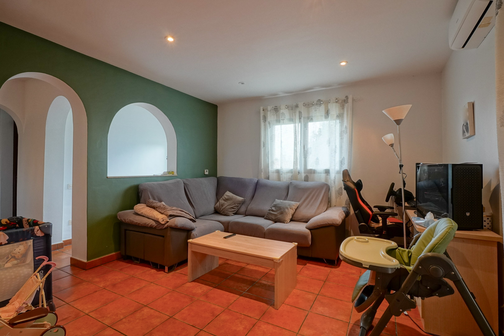 Living area featuring terracotta tile floors, a green accent wall with arched openings, and a grey sectional sofa. Equipped with split-system air conditioning.