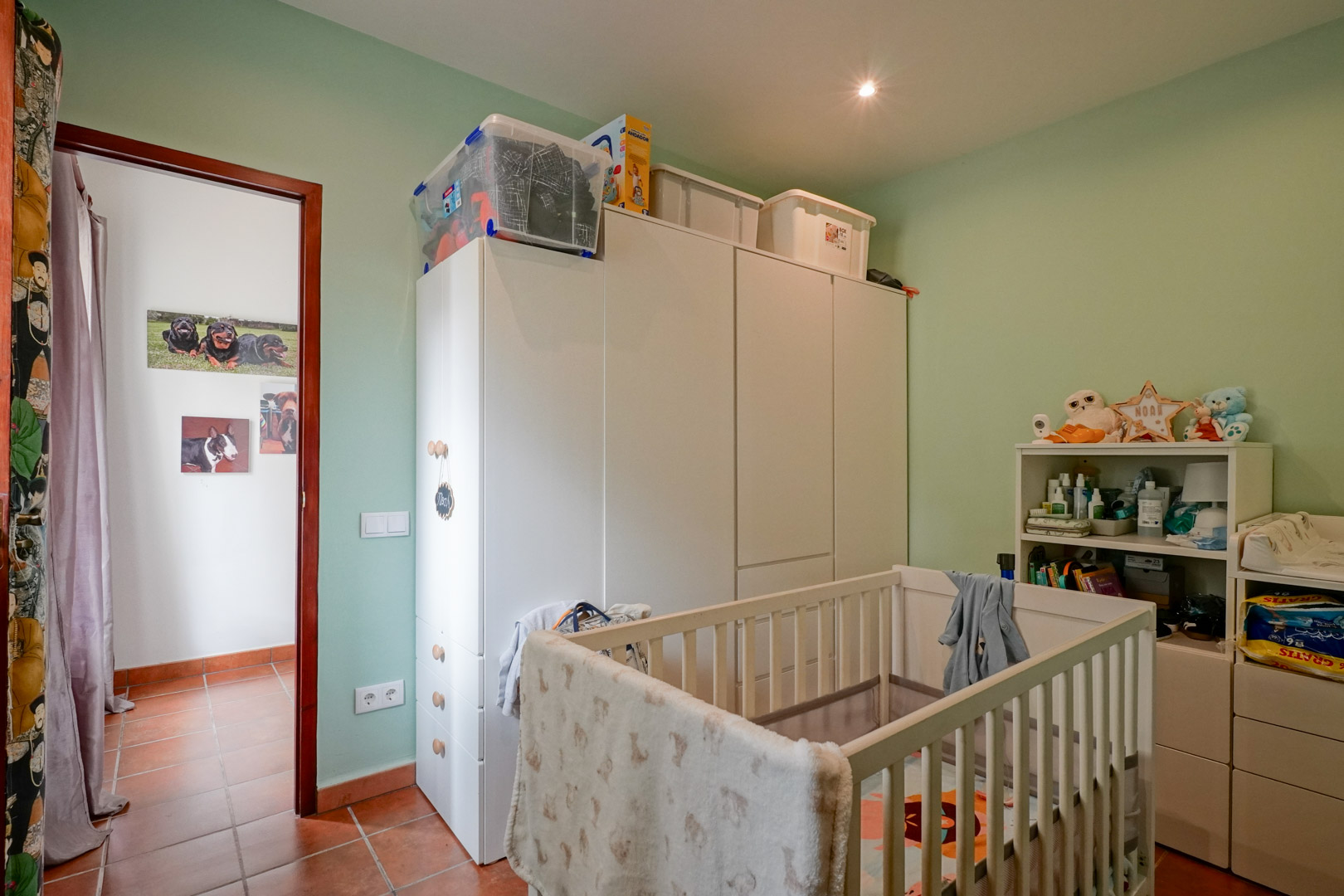 Nursery featuring a white wooden crib, matching storage wardrobes, and soft green walls. The space includes traditional terracotta tile flooring and recessed ceiling lighting.