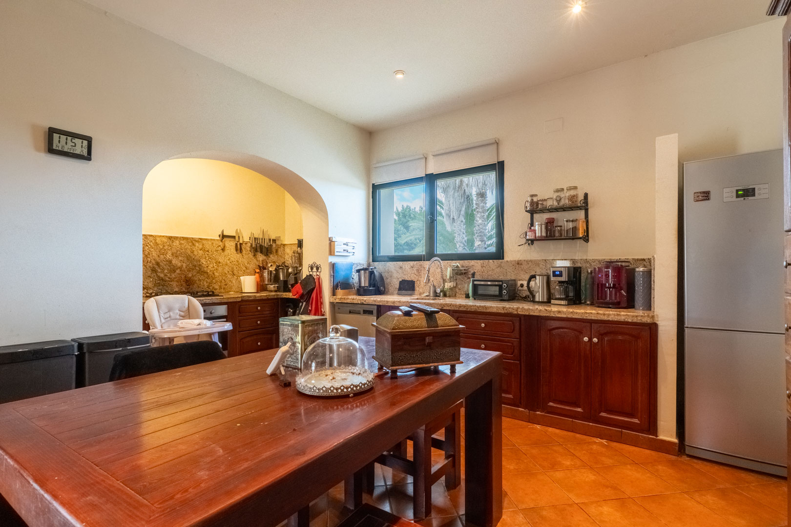Rustic kitchen featuring dark wood cabinetry, granite countertops, and a central dining table. An archway leads to the cooking zone, set on traditional terracotta tile flooring.