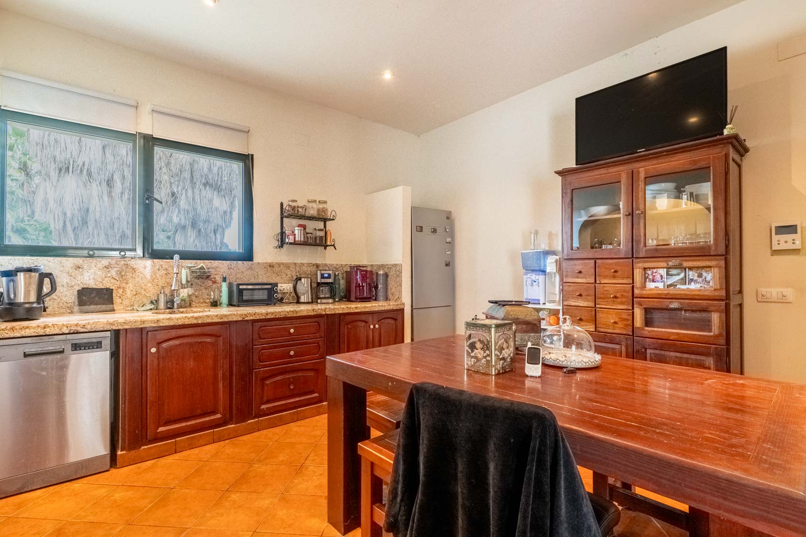 Rustic kitchen featuring granite countertops, dark wood cabinetry, and a large central dining table. Includes stainless steel appliances and natural light through the large window.