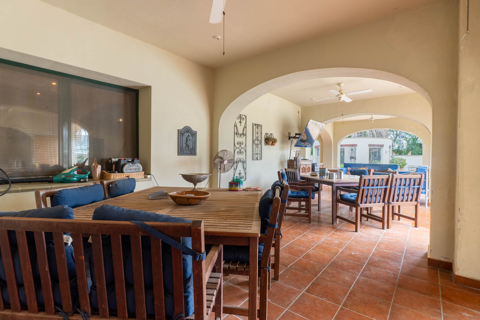 Spacious covered terrace featuring traditional Mediterranean arches, terracotta flooring, and wooden dining sets. Equipped with ceiling fans and views of the surrounding estate.