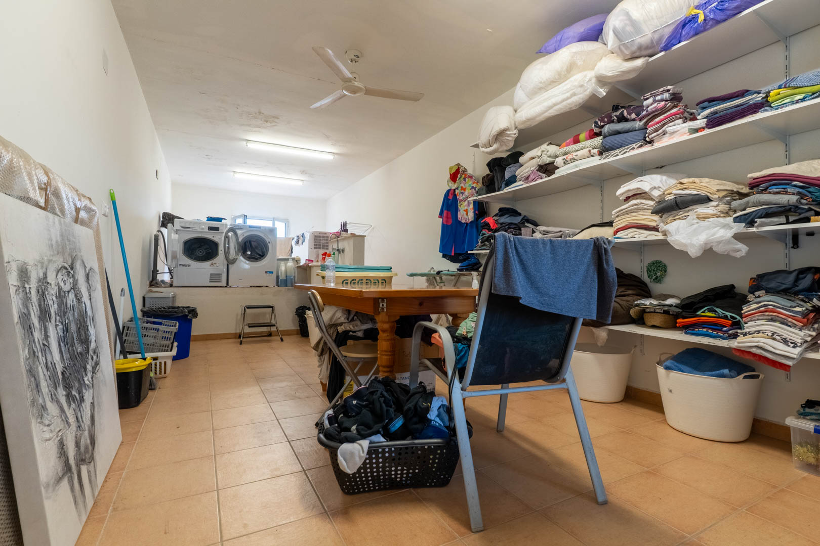 Spacious laundry and storage room featuring elevated washing machines, extensive open shelving for linens, and a central wooden work table.