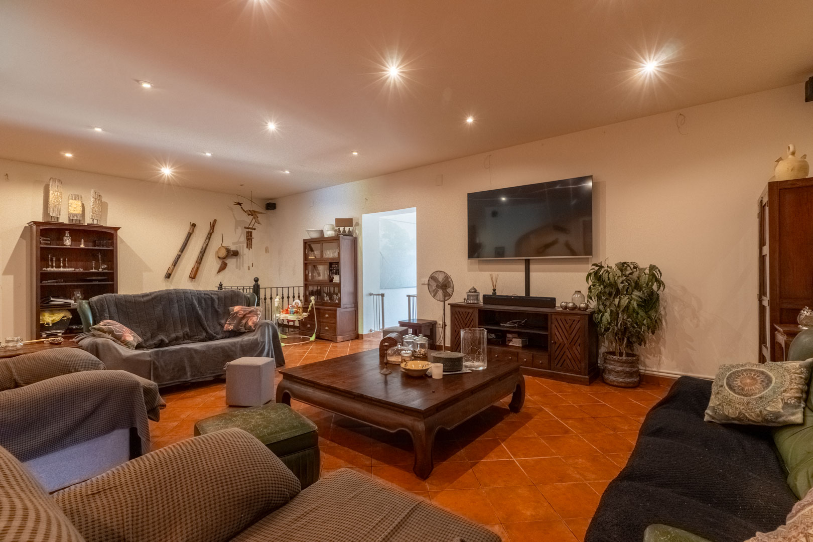 Spacious living area featuring rustic terracotta flooring, recessed ceiling spotlights, and solid wood furniture. The layout includes a media zone and access to the staircase.