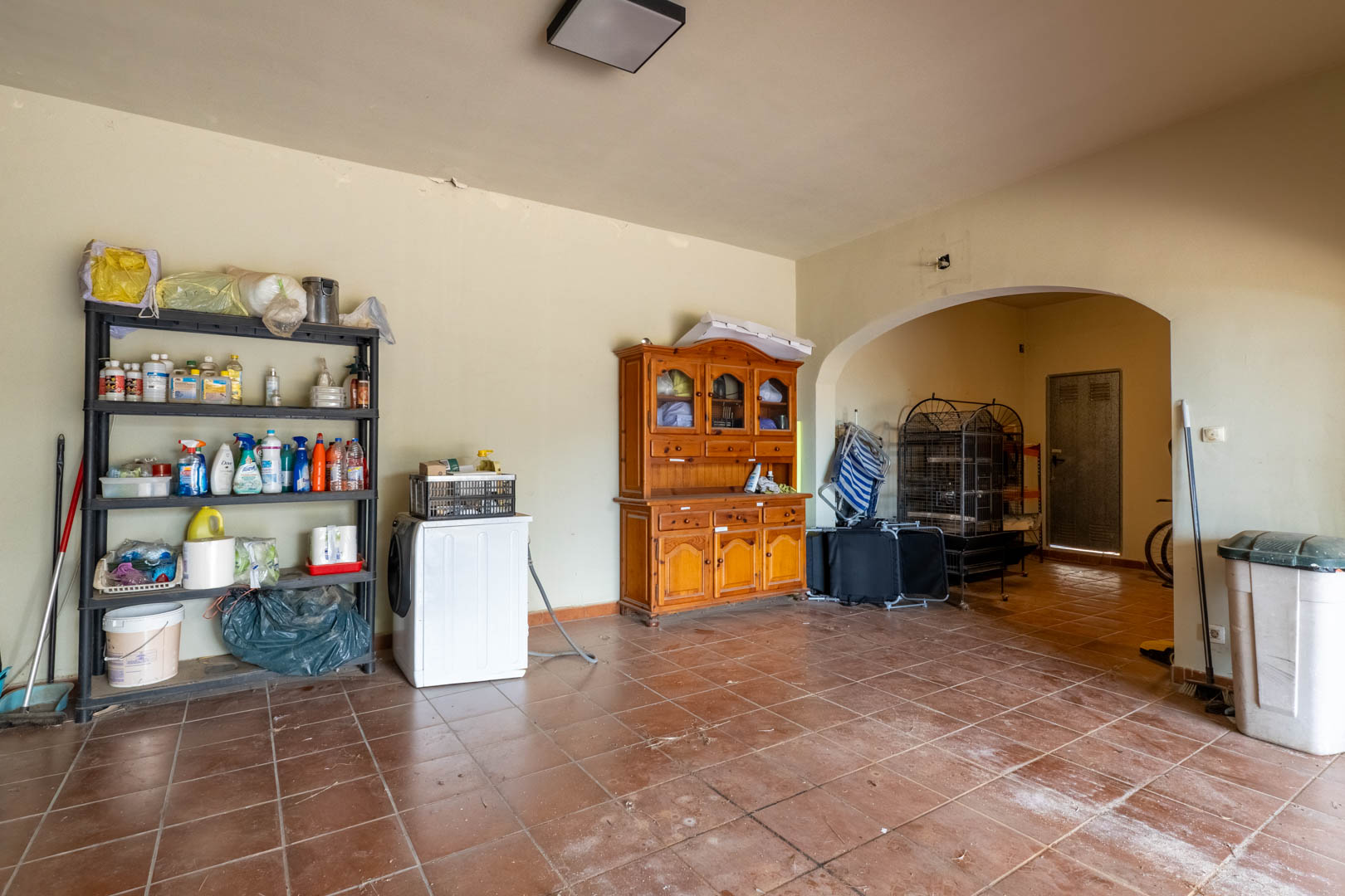 Spacious lower-level storage area featuring terracotta tile flooring, functional shelving, and an arched passage leading to the utility and garage zones.