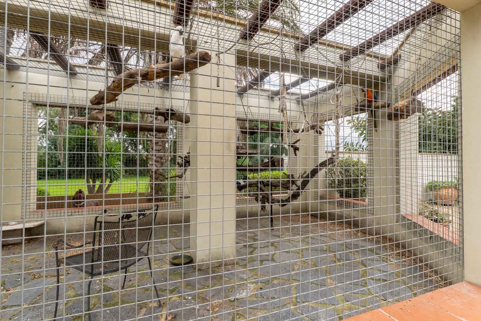 Spacious outdoor aviary featuring metal mesh fencing, slate flooring, and natural wood perches, set within the estate's landscaped gardens.