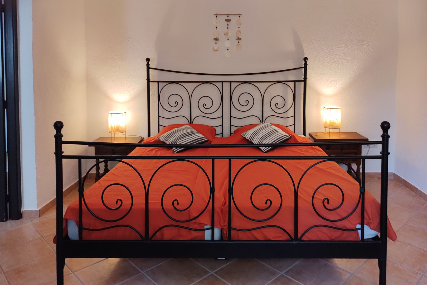 Bedroom featuring a black wrought iron bed frame with scrollwork, wooden nightstands, and ceramic tile flooring. Warm bedside lamps illuminate the textured white walls.