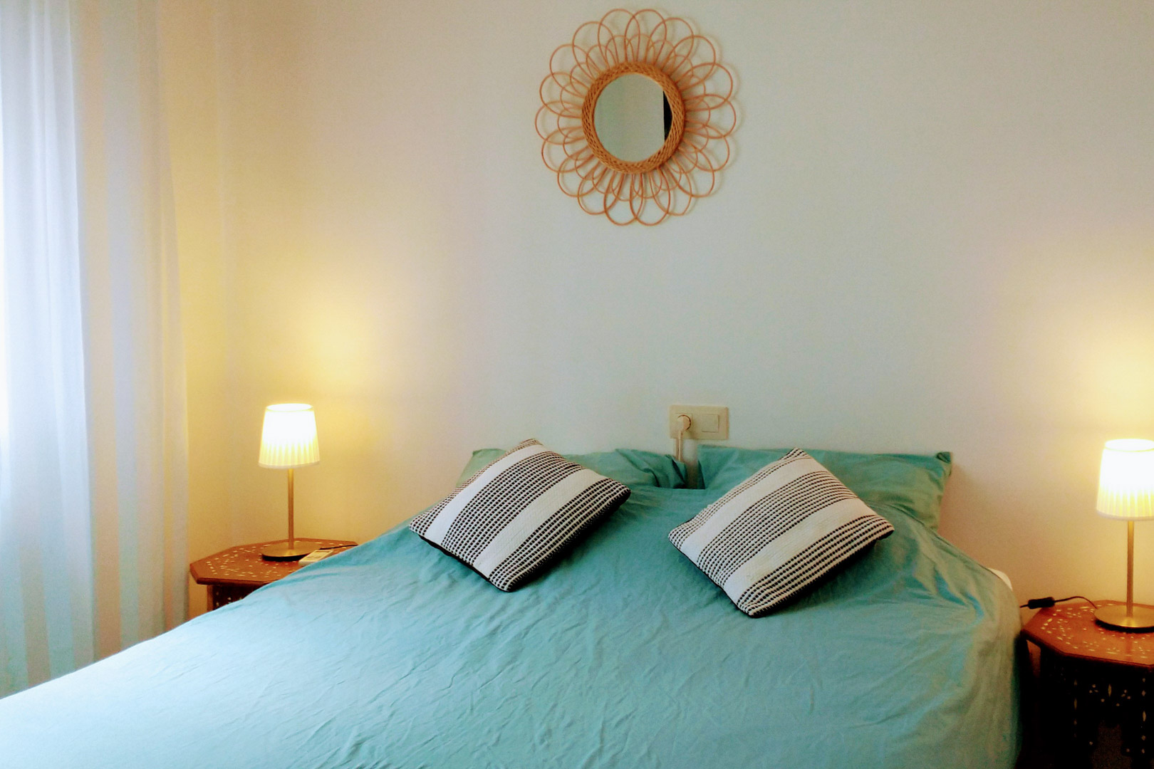 Bedroom featuring a double bed with turquoise linens, wooden hexagonal side tables with warm lighting, and a decorative rattan sunburst mirror.