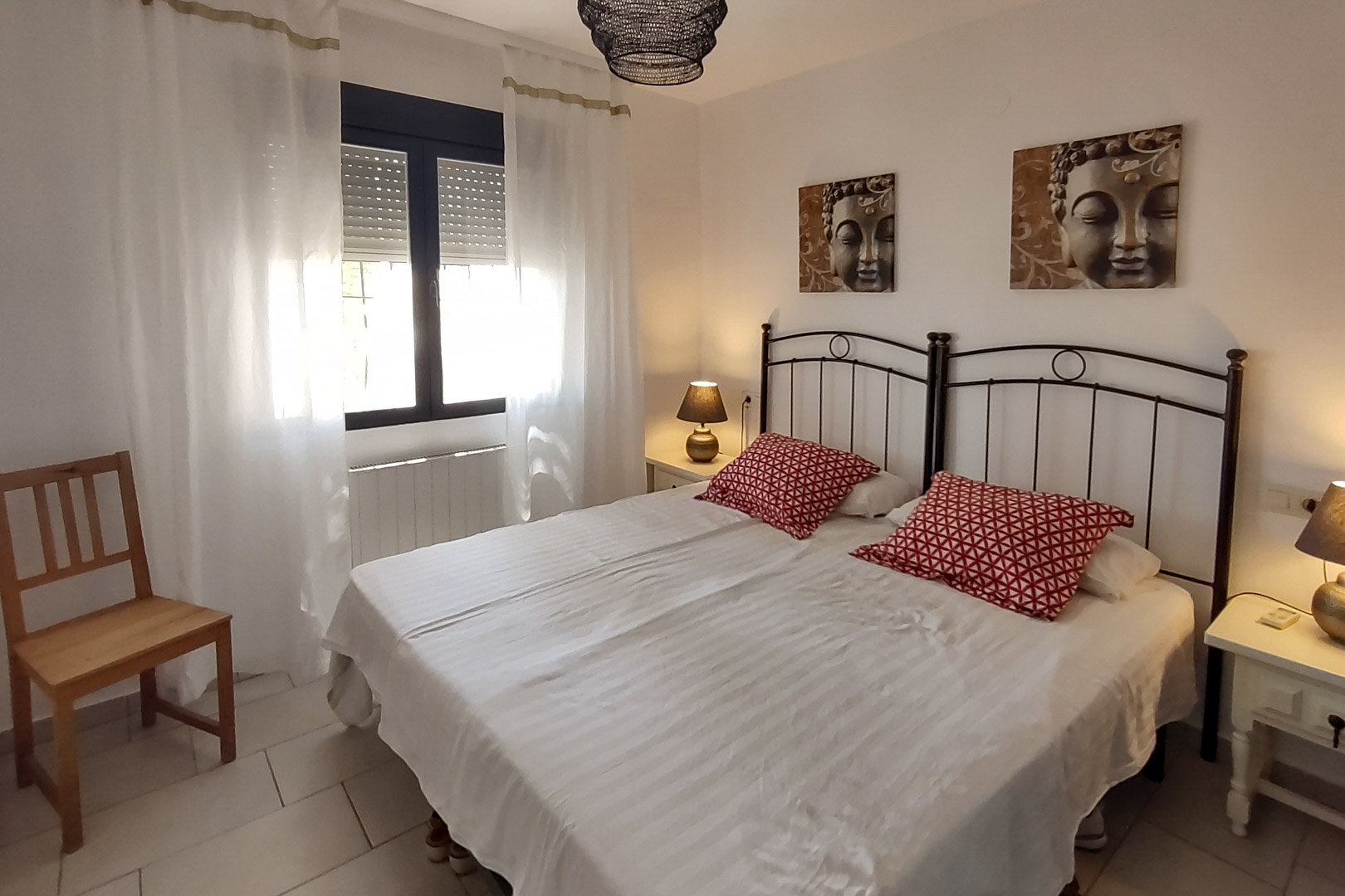 Bedroom featuring twin beds with black wrought iron headboards, Buddha artwork, and a window equipped with roller shutters and white curtains.