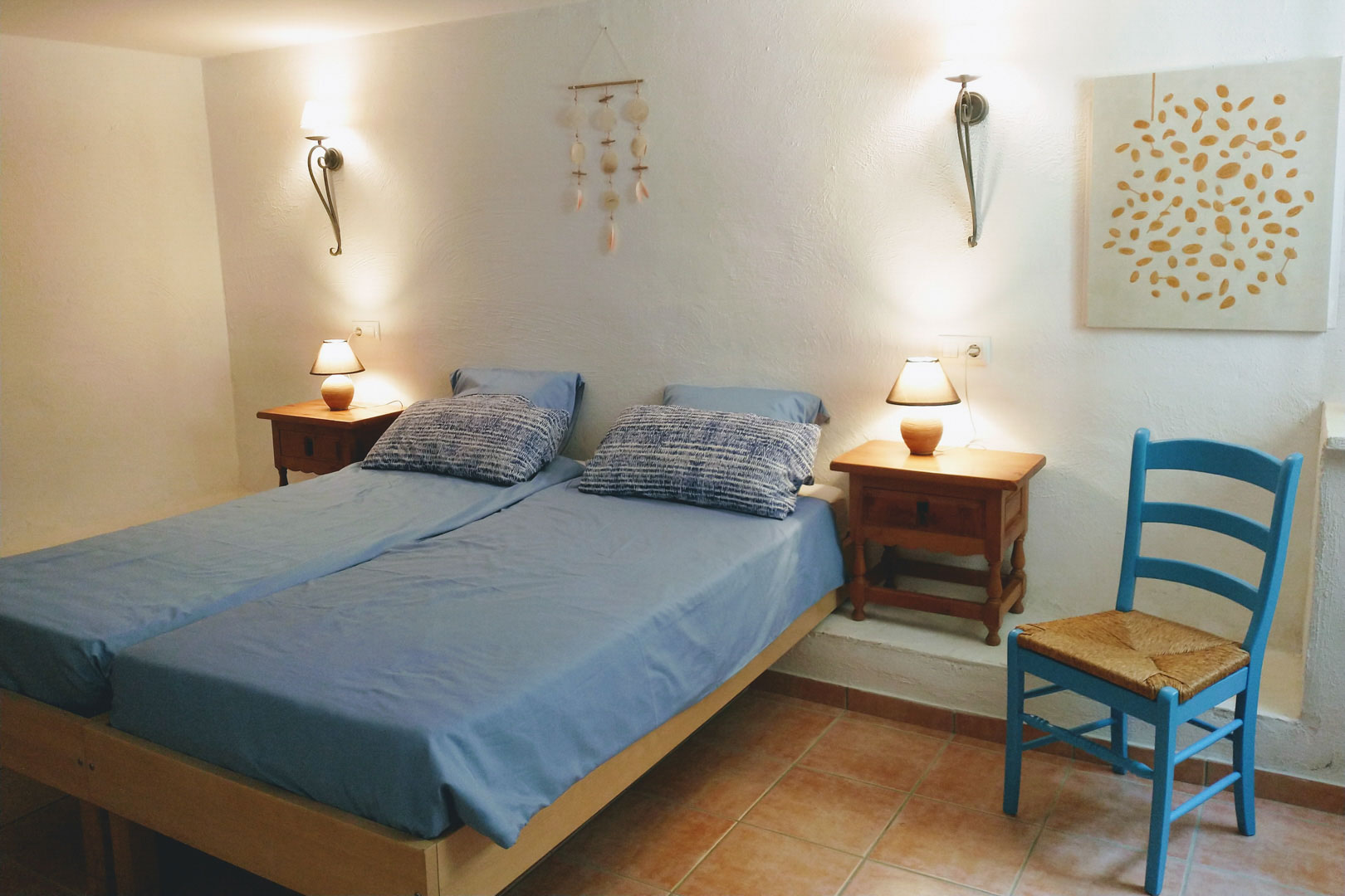 Bedroom featuring twin beds with blue linens, terracotta tile flooring, and rustic wooden nightstands. Warm lighting is provided by wall sconces and table lamps.