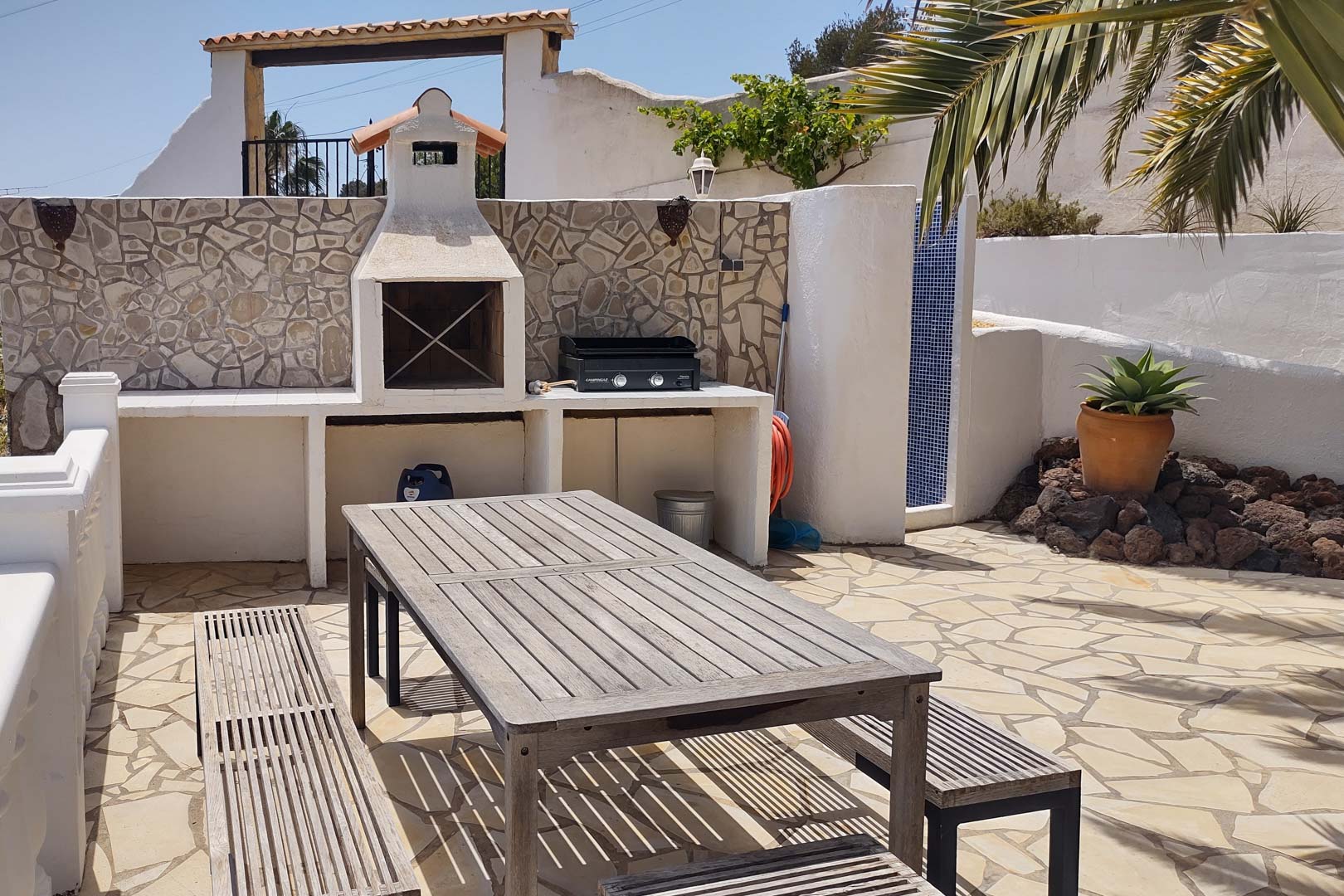 Built-in masonry barbecue and gas grill area featuring a wooden picnic table, natural stone paving, and a nearby outdoor shower with blue mosaic tiles.