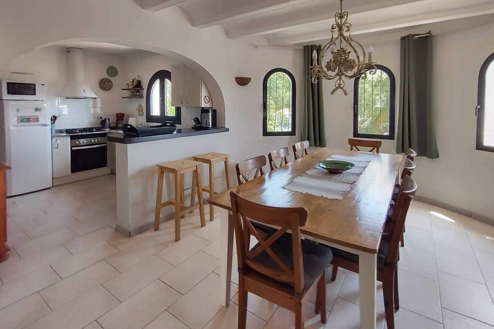 Dining area featuring a large wooden table for eight, connected via an archway to a kitchen with a breakfast bar, stools, and Mediterranean arched windows.