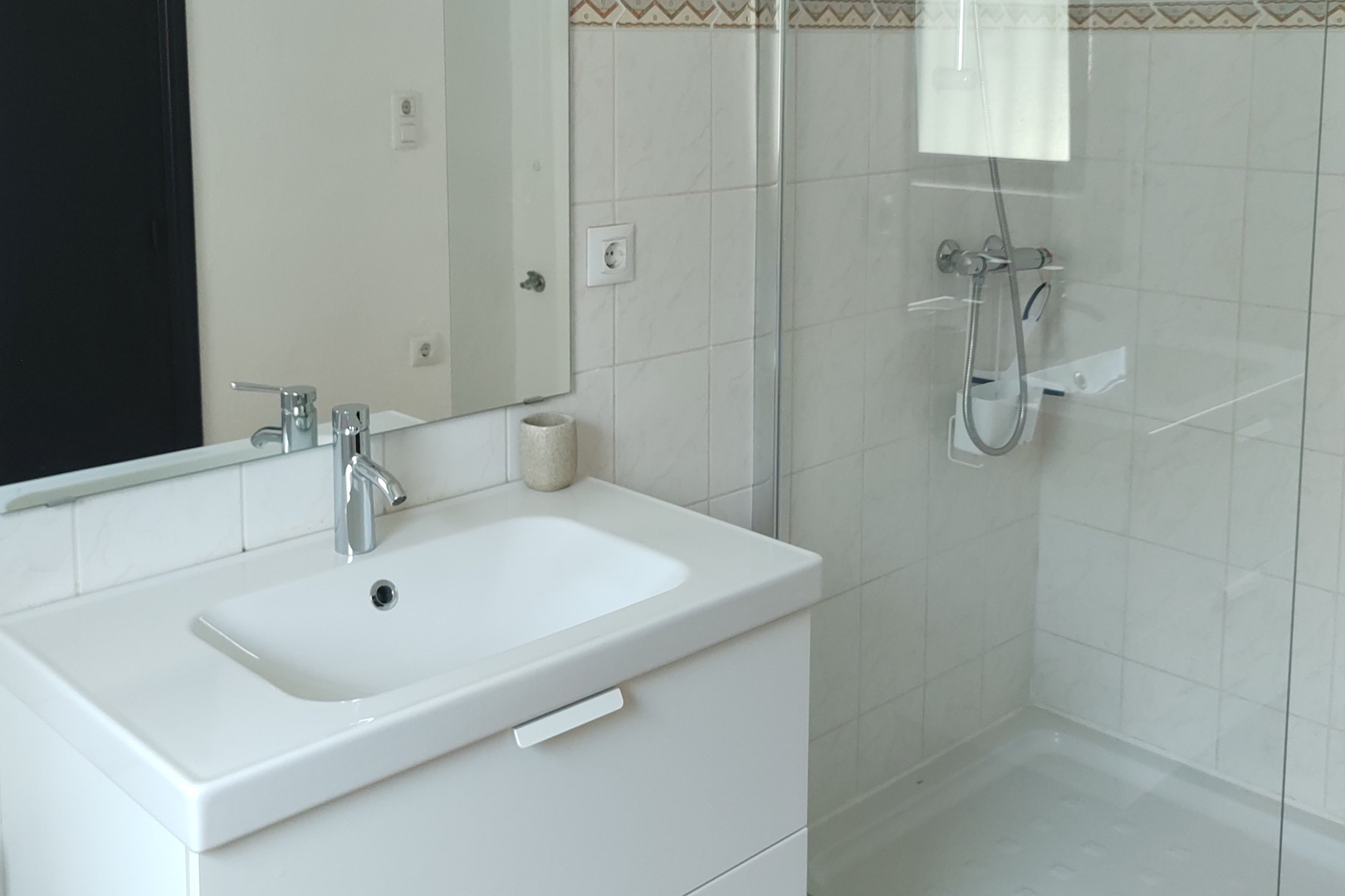 Functional bathroom featuring a white vanity unit with chrome fixtures and a walk-in shower with a glass screen. The walls are finished with white tiles and a decorative border.