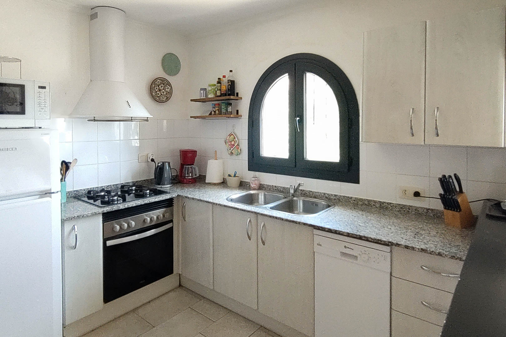 Functional kitchen featuring granite countertops, a gas hob, and white cabinetry. An arched dark-framed window provides natural light over the double stainless steel sink.