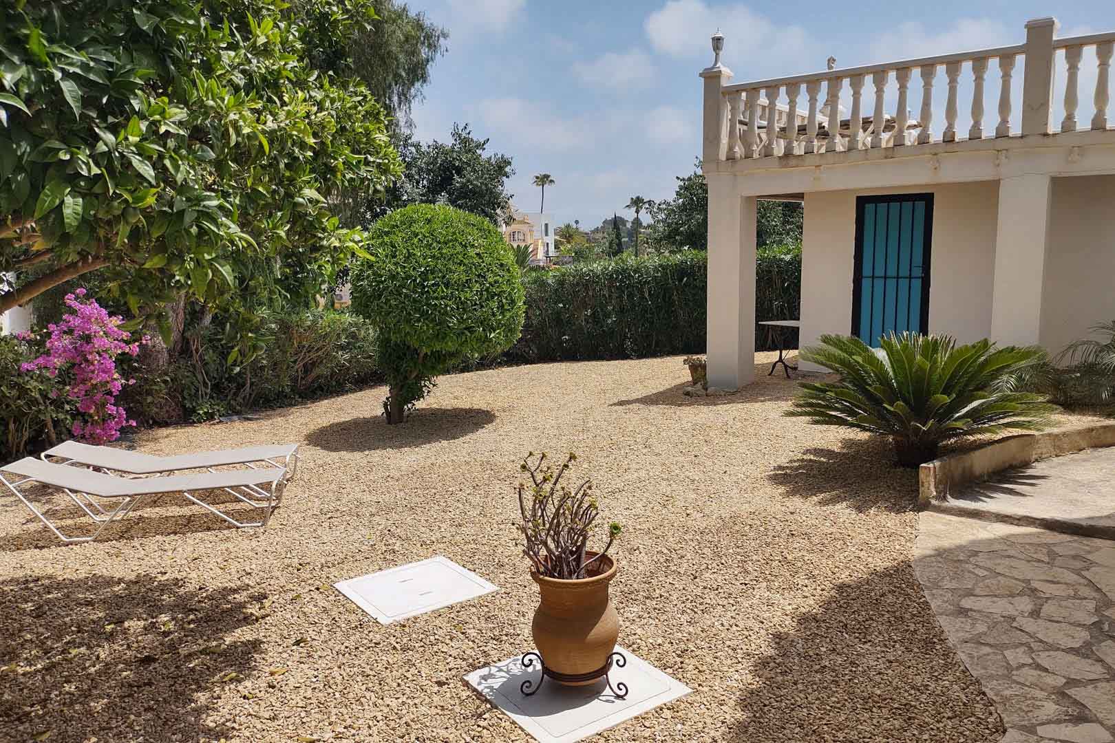 Low-maintenance Mediterranean garden featuring a gravel area, sun loungers, native plants, and a white facade with a classic stone balustrade.