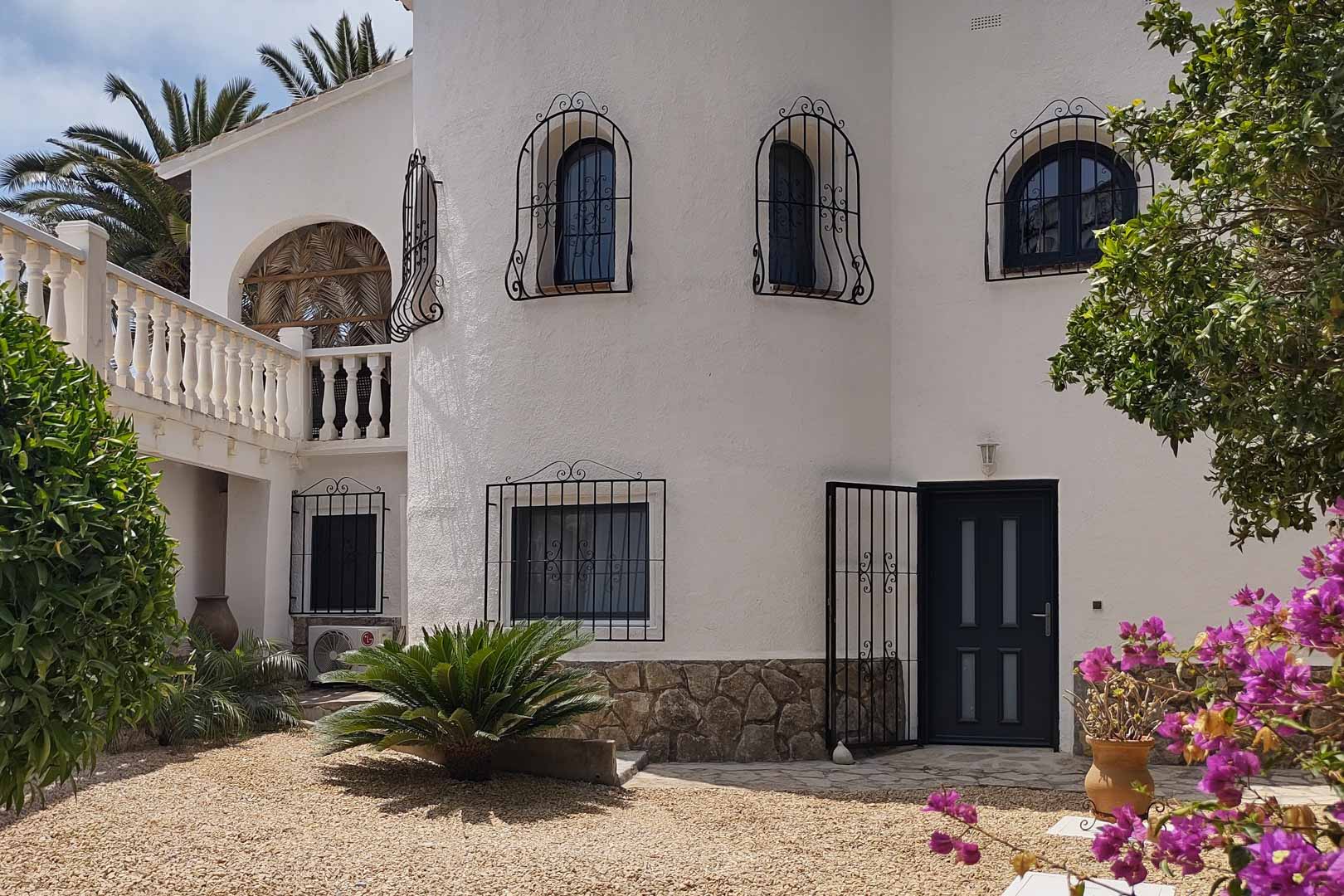 Mediterranean villa exterior featuring white walls, stone accents, decorative wrought iron window grilles, and a gravel garden entrance framed by vibrant bougainvillea.