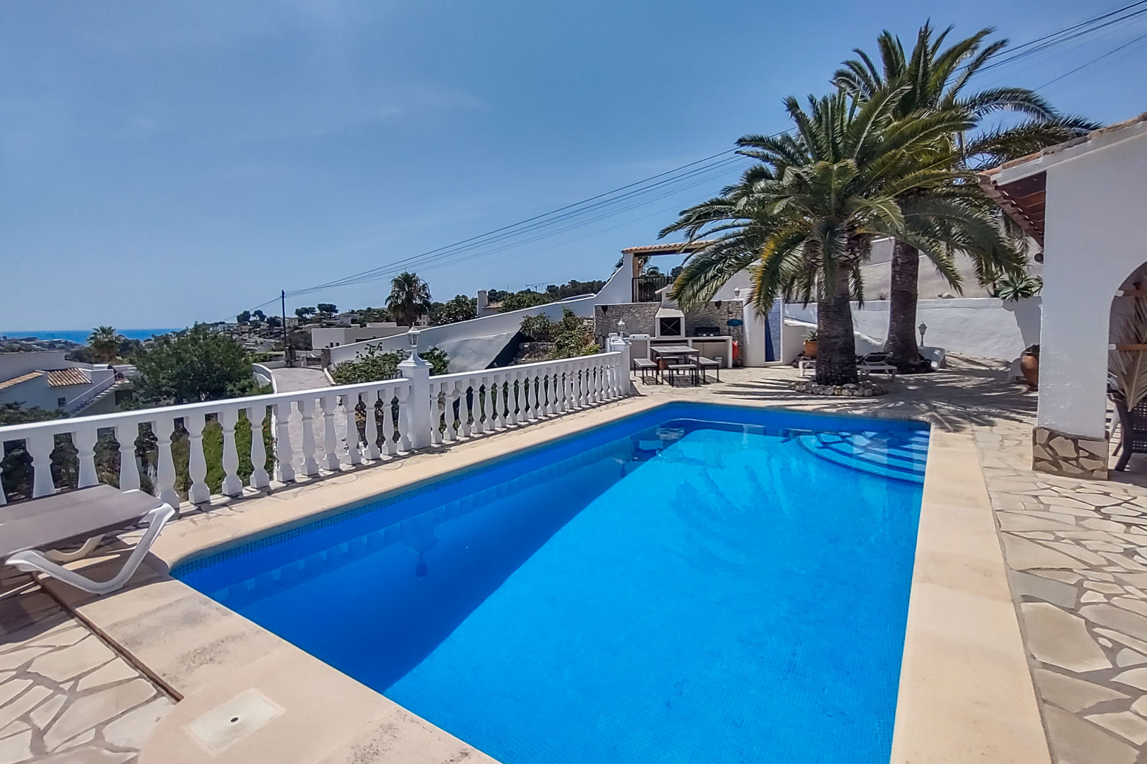 Outdoor pool area featuring a stone-paved terrace, white balustrade, and sea views. Includes a background BBQ zone with palm trees and a picnic area.