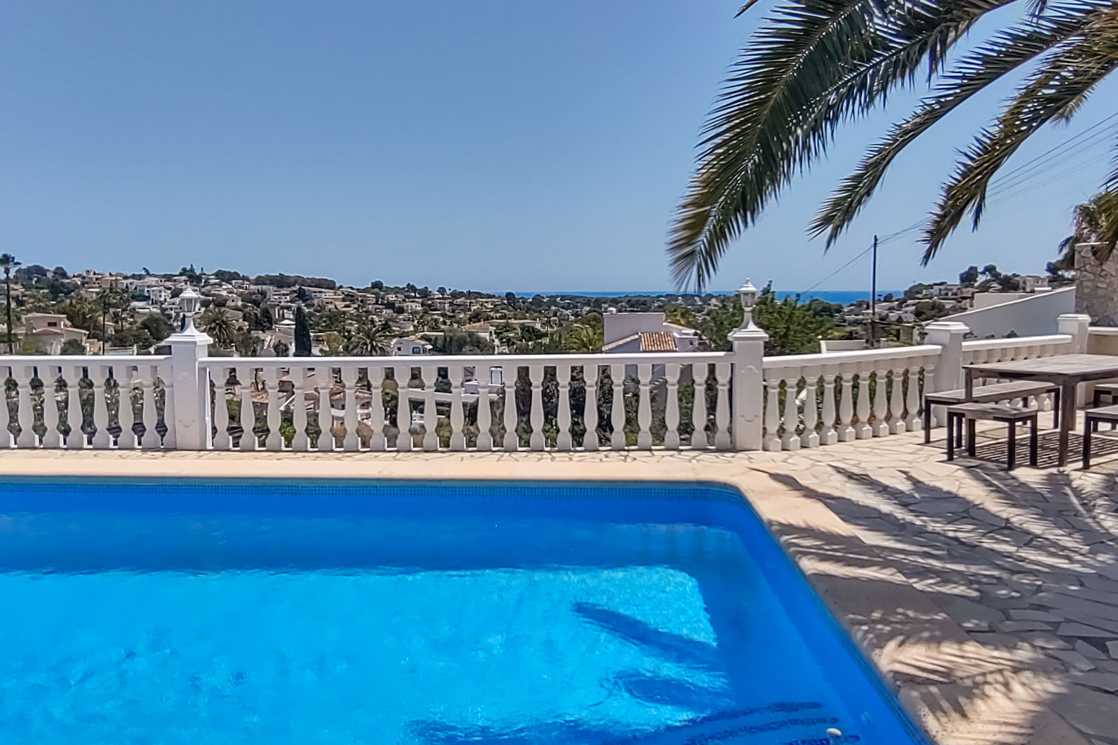 Pool terrace with a white balustrade overlooking the Mediterranean Sea and valley, featuring natural stone paving and a wooden outdoor dining set.