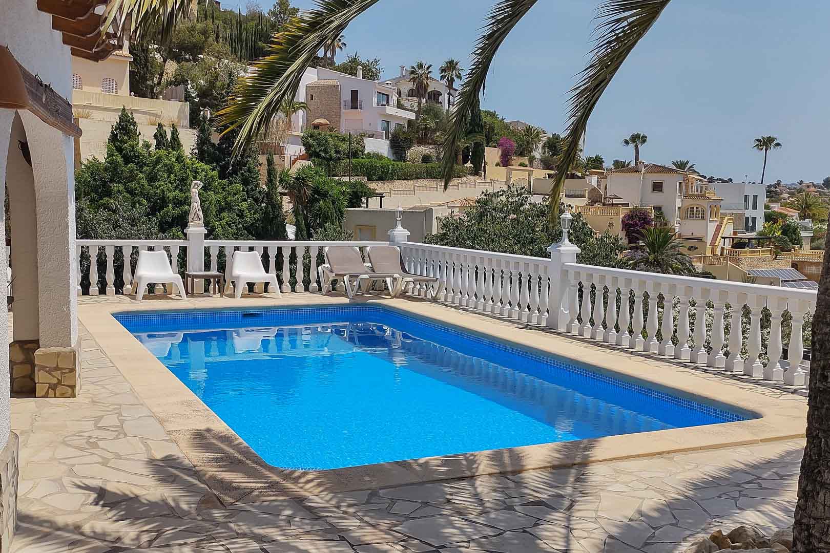 Rectangular pool with blue mosaic tiles, set within a natural stone terrace and classic white balustrade, offering elevated views of the coastal hillside.
