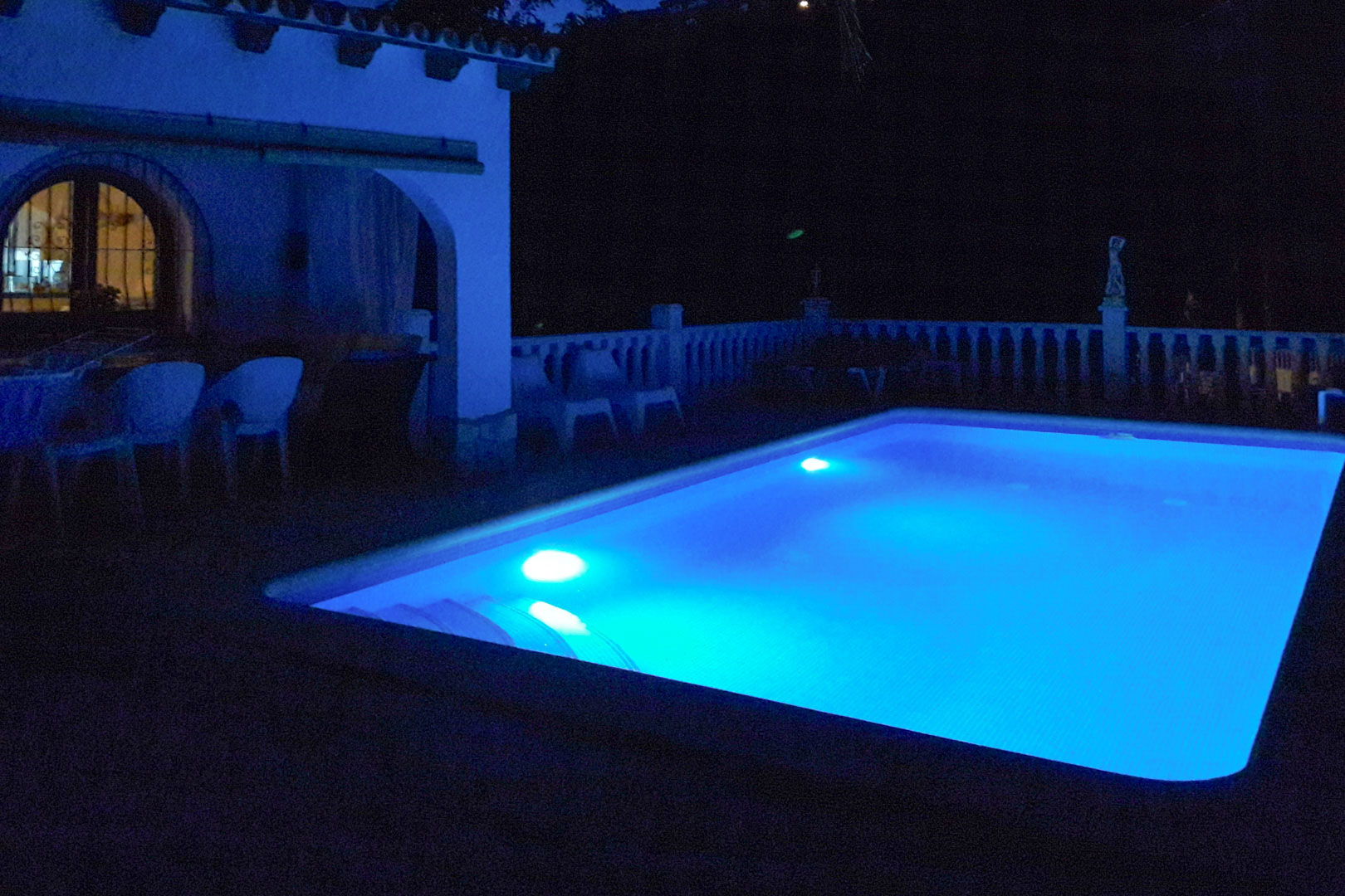 Rectangular swimming pool featuring vibrant blue underwater LED lighting, set beside an arched terrace with outdoor seating and a white balustrade.
