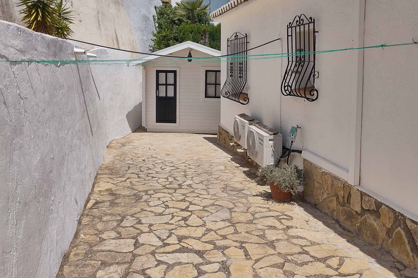 Side path with natural stone paving leading to a wooden garden shed, featuring exterior air conditioning units and traditional wrought iron window security bars.