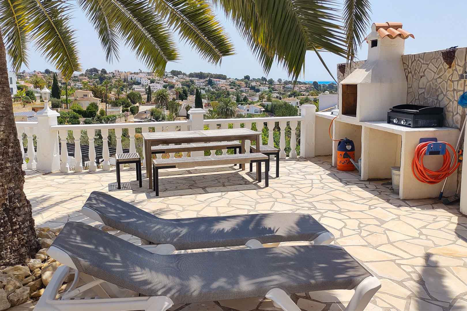 Stone-paved outdoor terrace featuring a built-in barbecue station, wooden picnic seating, and sun loungers with panoramic valley and sea views.