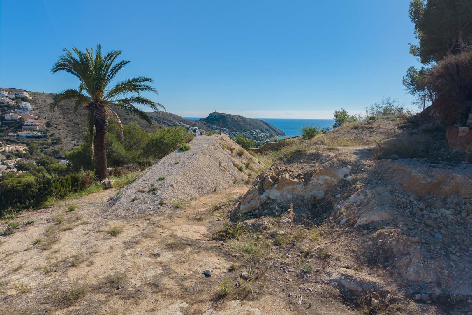 Elevated building plot under preparation featuring panoramic views of the Mediterranean Sea and Moraira coastline, framed by natural vegetation and palm trees.