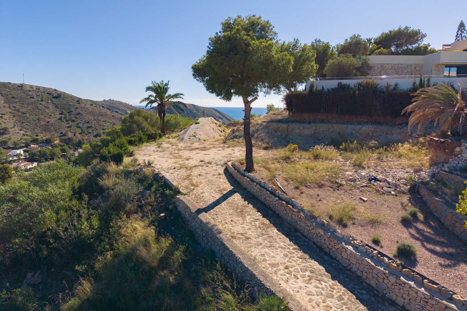 Panoramic view from the plot featuring a stone-paved path and dry stone walls, surrounded by Mediterranean flora with clear sea views.
