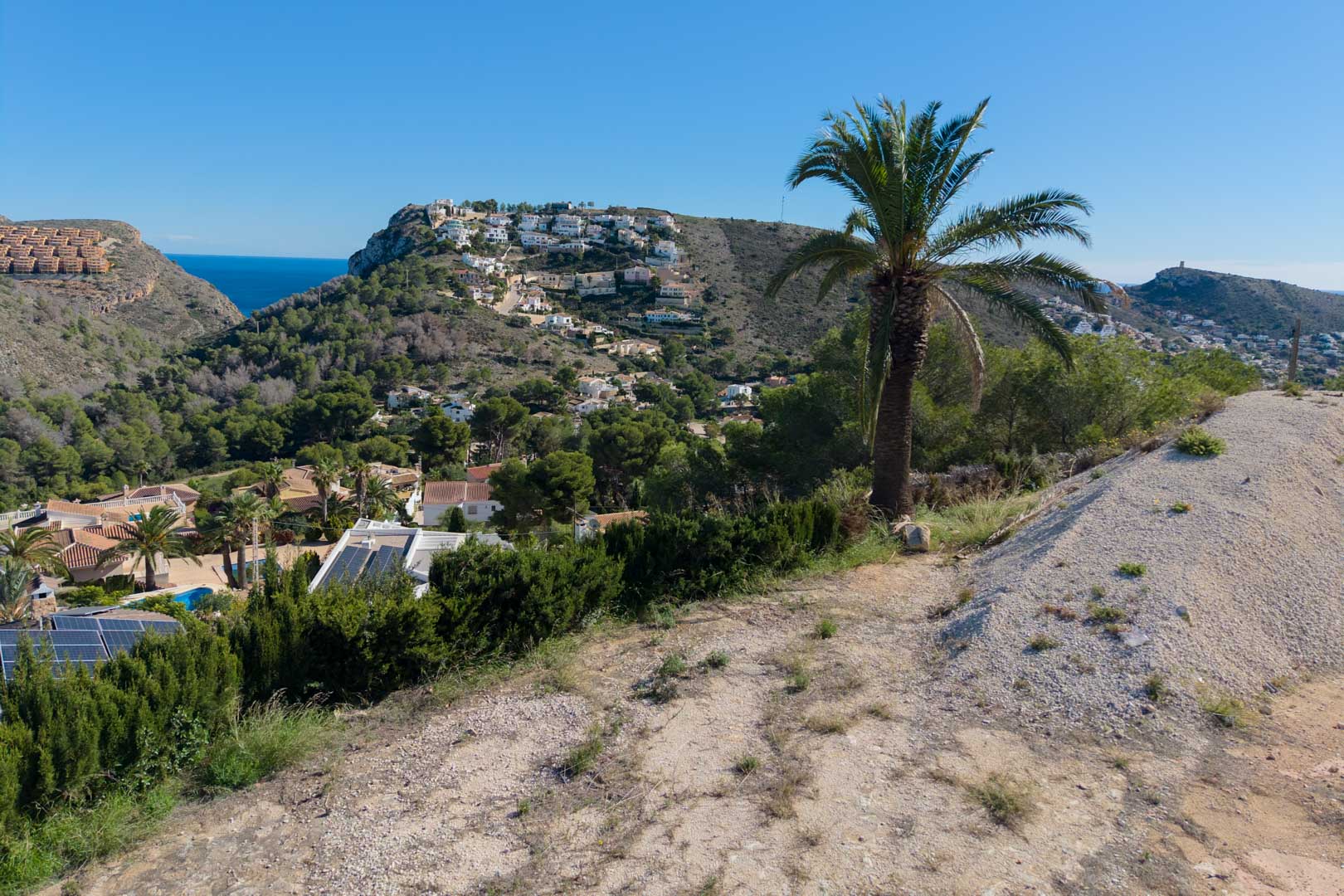Panoramic views from the elevated plot overlooking the Moraira valley, featuring the Mediterranean Sea and hillsides with lush pine and palm trees.