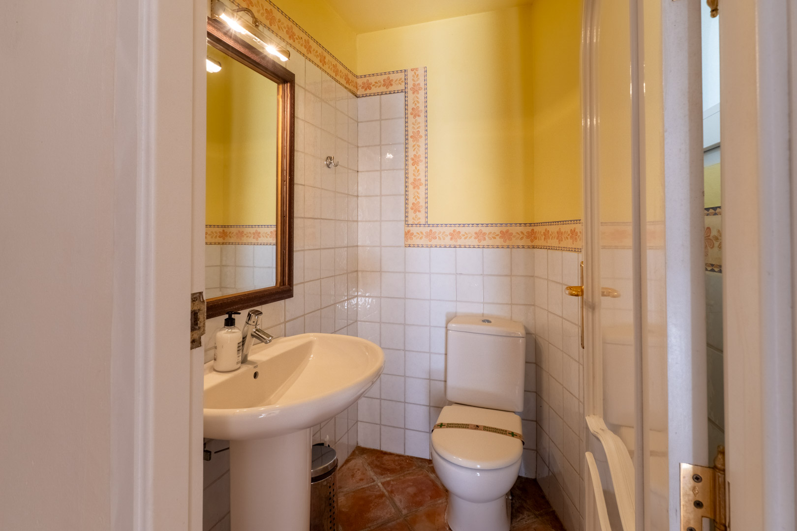 Guest powder room featuring terracotta flooring, white wall tiles with decorative floral borders, and a pedestal sink beneath a wood-framed mirror.