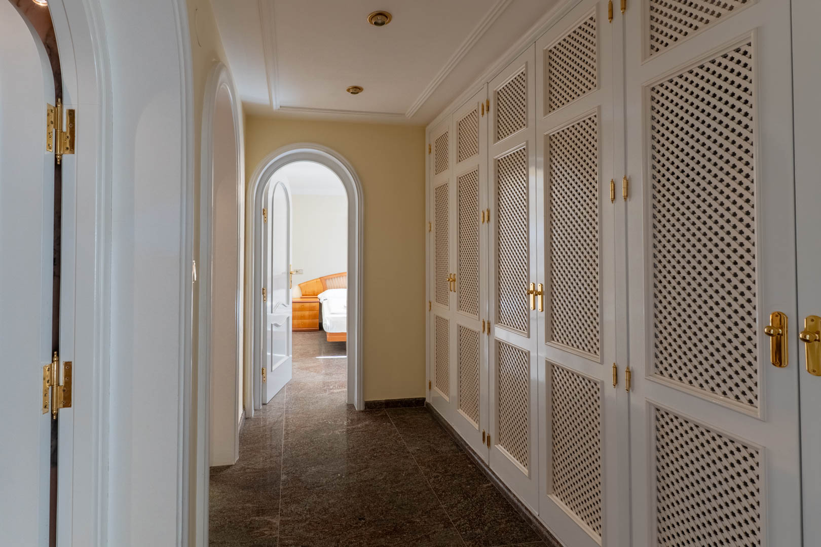 Hallway featuring white lattice-door built-in wardrobes with gold hardware, dark granite flooring, and arched doorways leading to the bedrooms.