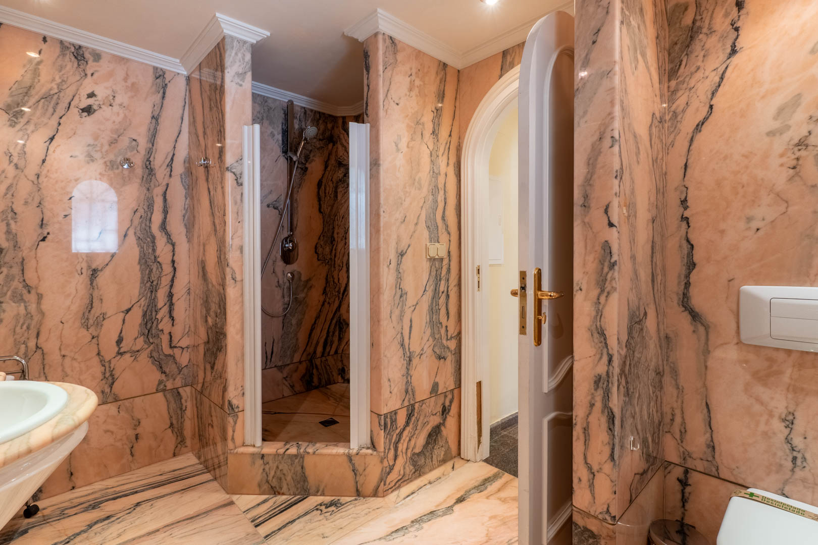Luxury bathroom featuring full pink marble wall and floor tiling, a walk-in shower, classic crown moldings, and an arched doorway with gold-toned hardware.
