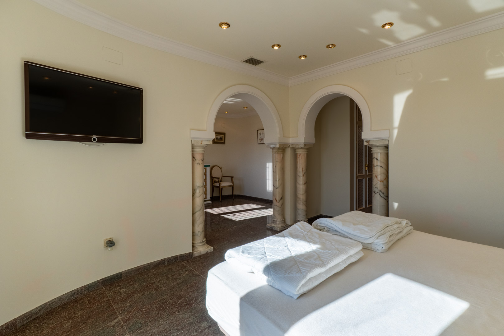 Master bedroom featuring classical arches with marble columns, polished stone flooring, and a wall-mounted TV leading to an adjoining private area.