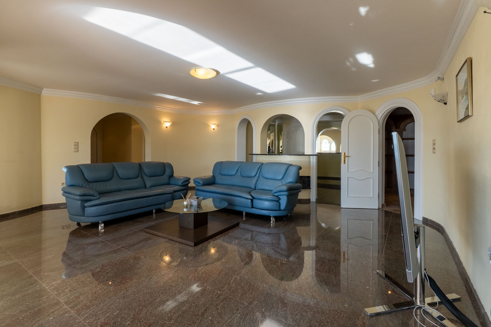 Spacious lounge with reflective marble flooring, blue leather seating, and architectural arches leading to a bar area and adjacent rooms.