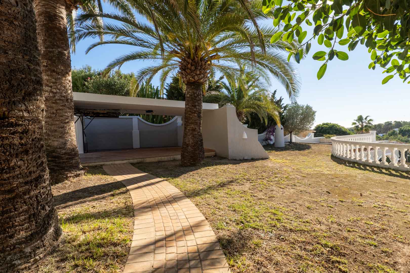 Stone-paved path leading to a covered carport, framed by mature palm trees and a classic white balustrade overlooking the landscape.