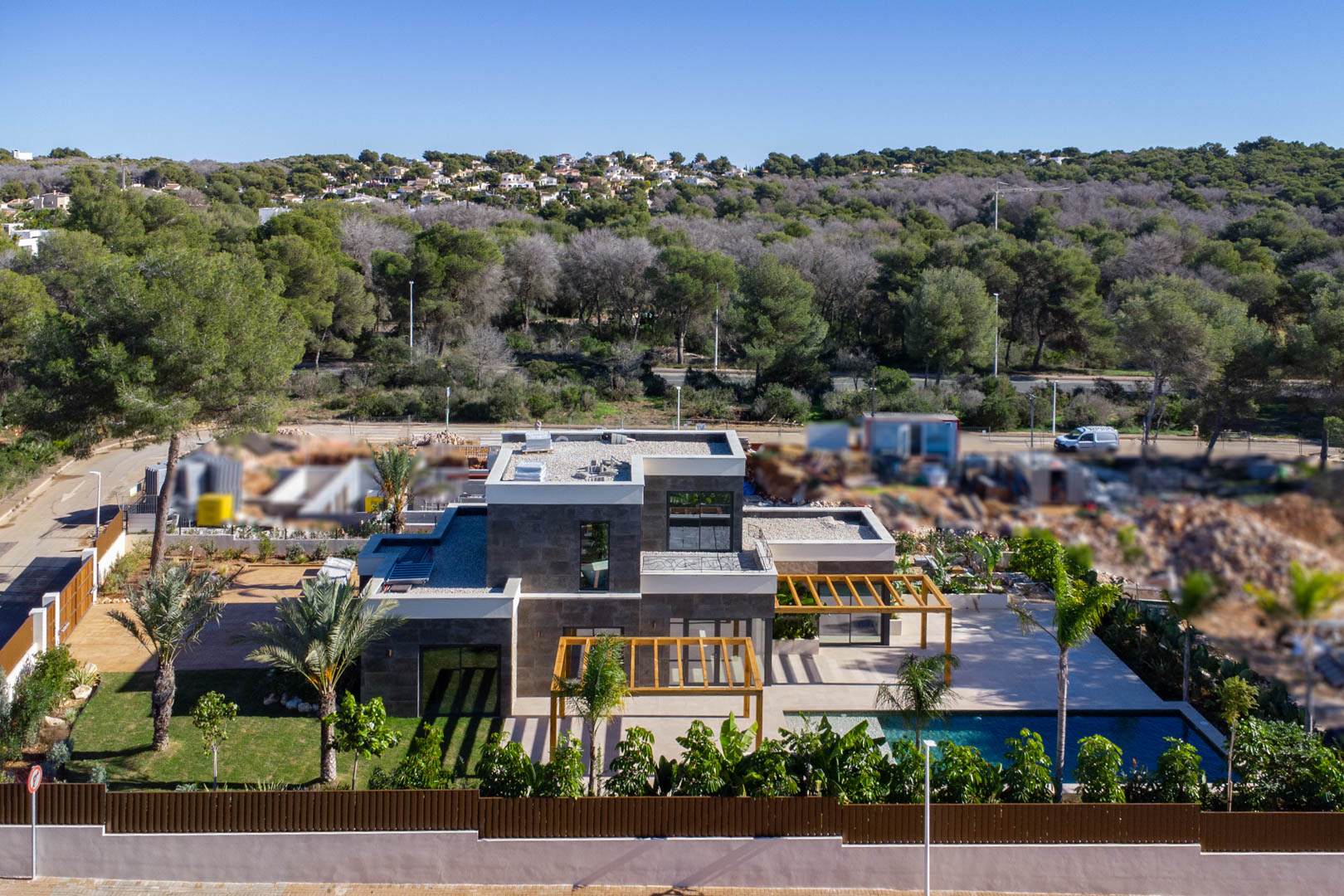 Aerial view of the modern villa featuring stone cladding, iroko wood pergolas, a Bali-style pool, and landscaped gardens set against a lush green backdrop.