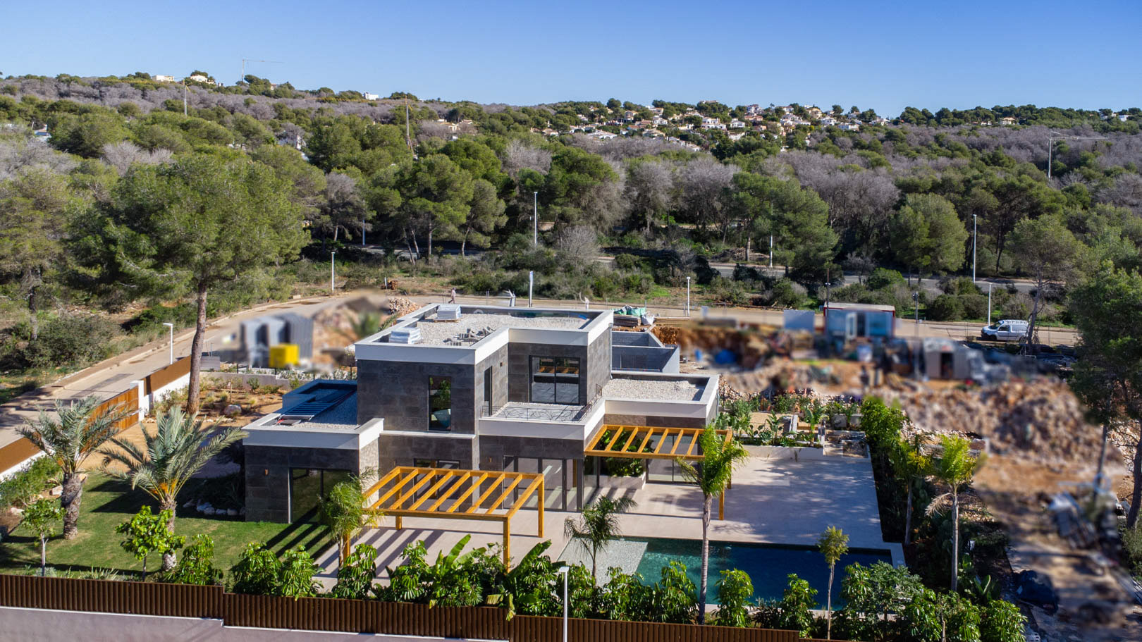 Aerial view of the modern villa with dark stone cladding, iroko wood pergolas, and a saltwater pool, set within a landscaped Mediterranean garden.