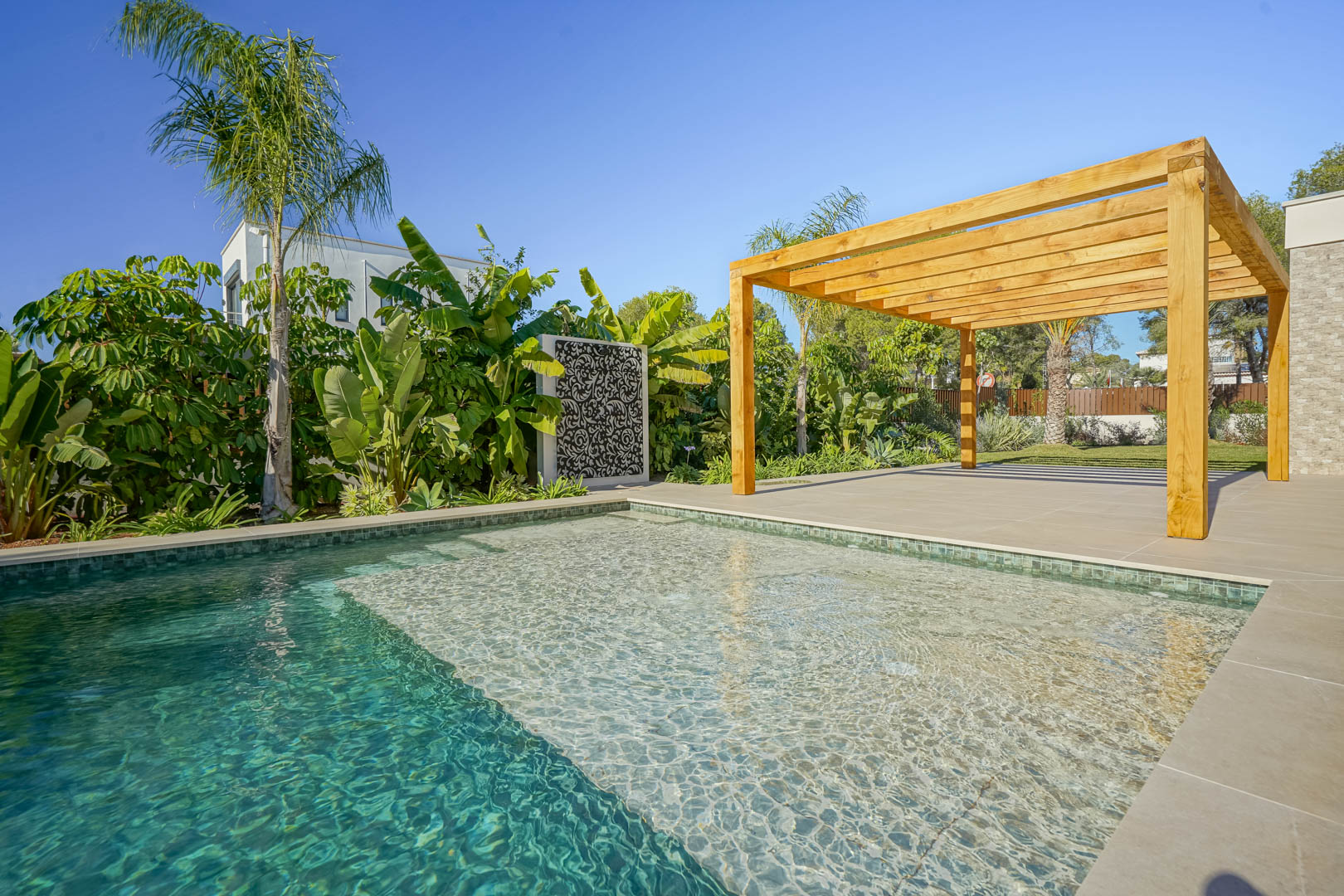 Bali-style saltwater pool featuring a shallow lounging area, adjacent to a large porcelain terrace with a wooden iroko pergola and lush landscaping.
