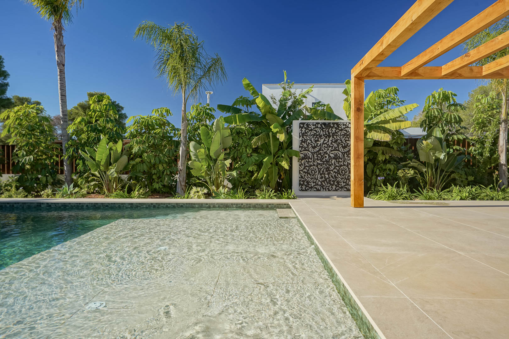 Bali-style saltwater pool featuring a shallow lounging area, light-toned terrace tiling, an iroko wood pergola, and a landscaped garden with tropical plants.