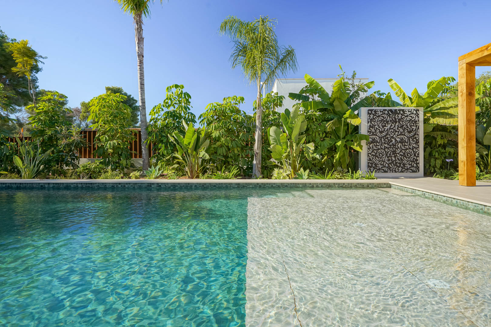 Bali-style saltwater pool with a shallow sun shelf, bordered by a lush Mediterranean garden featuring tropical plants and decorative screens.