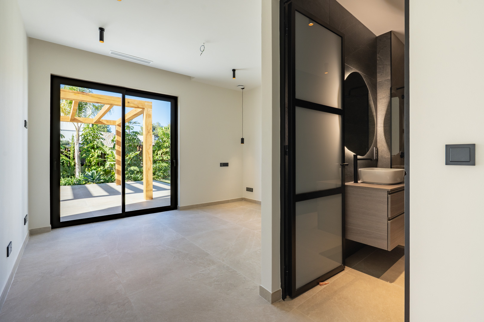 Bedroom with direct terrace access and a wooden pergola. Features an en-suite bathroom with a vessel sink, backlit mirror, and minimalist black fixtures.