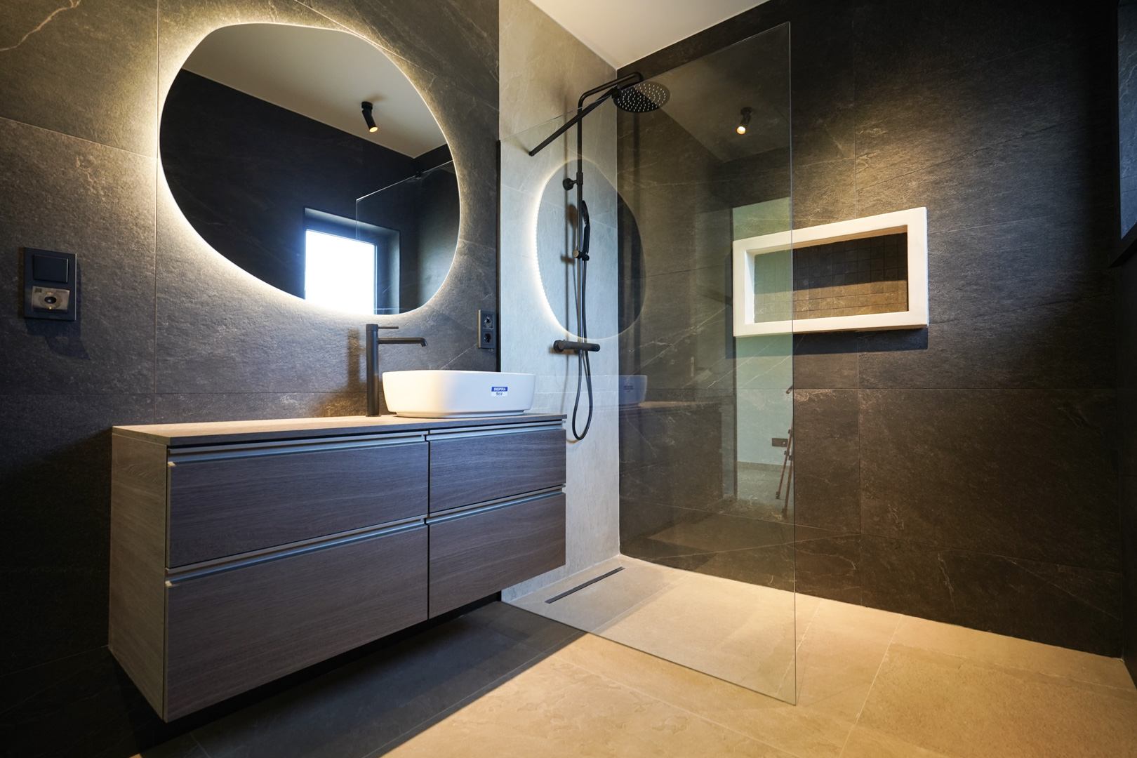 Designer bathroom with dark stone walls, a vessel sink, and a backlit organic mirror. Features a walk-in shower with black fixtures and a seamless glass partition.