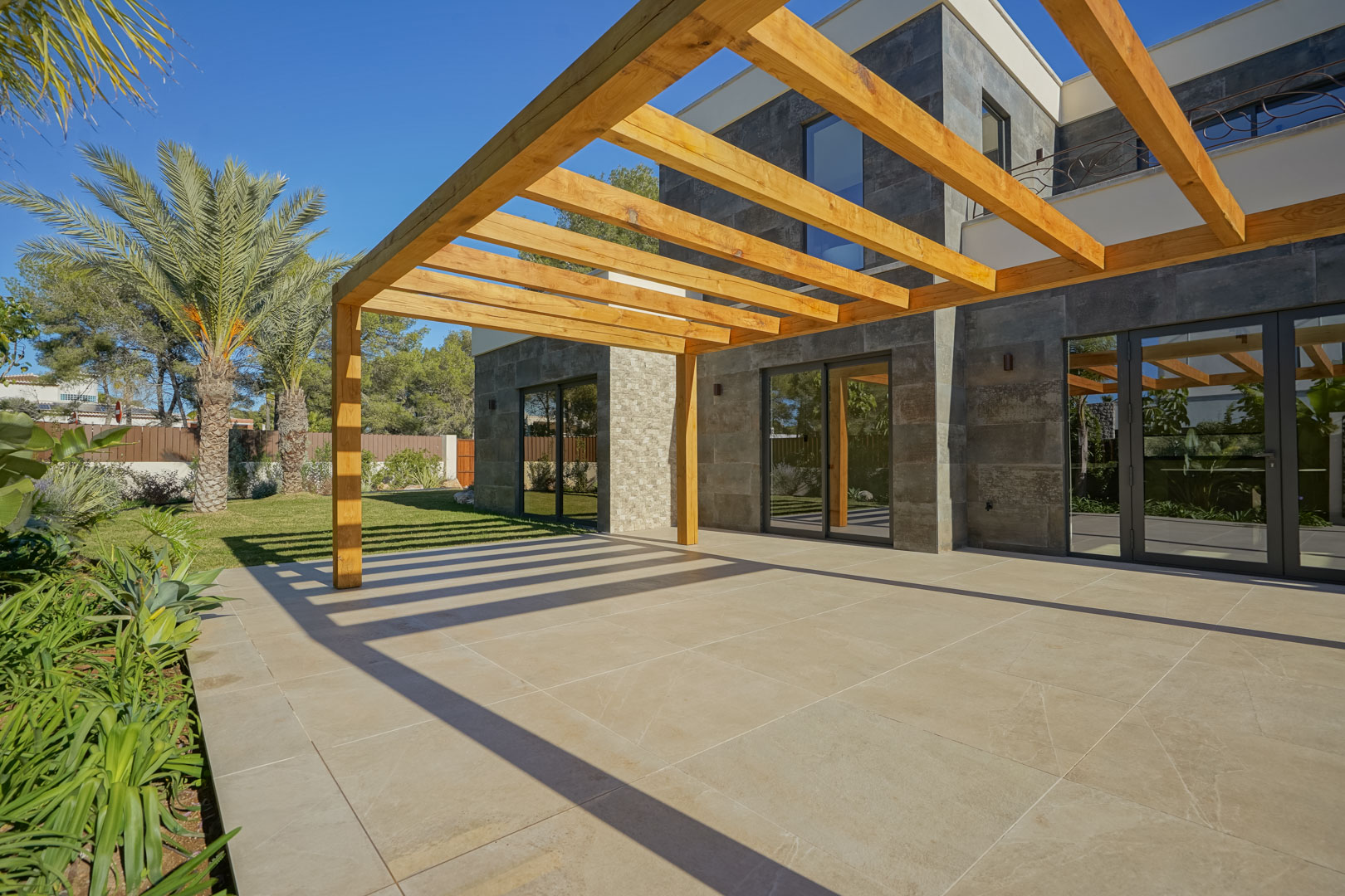 Exterior terrace featuring an iroko wood pergola and large-format tiling, set against a dark stone facade and landscaped Mediterranean garden.