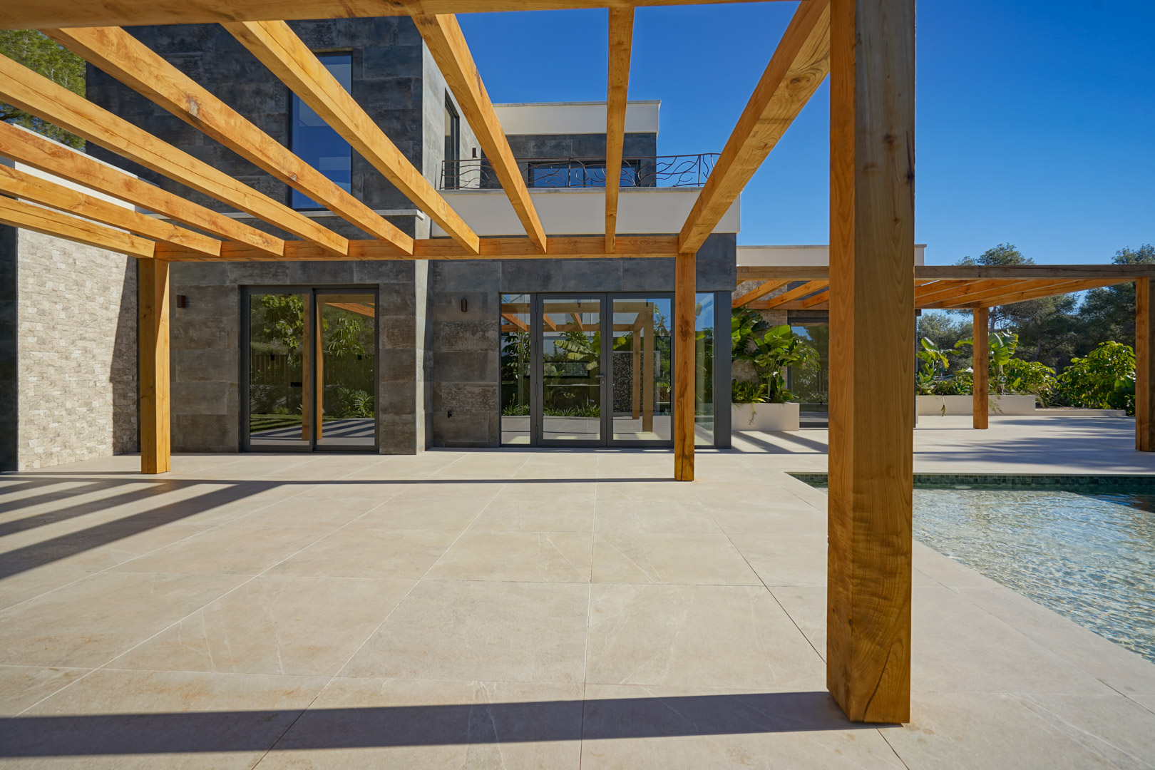 Exterior terrace featuring an iroko wood pergola and porcelain tiling, adjacent to the pool and a modern facade with large glass doors.